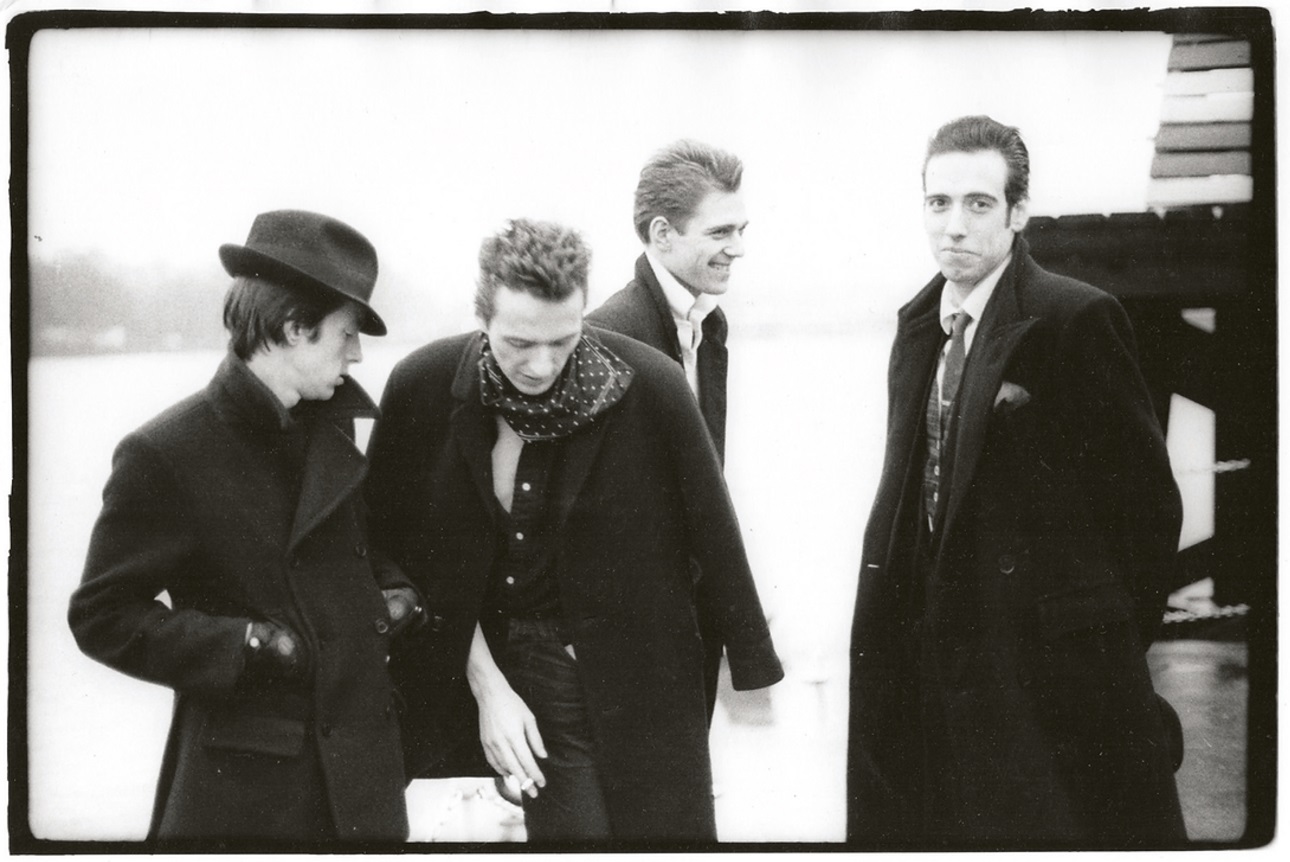 The Clash in a black and white group shot by Pennie Smith. They are in a row & smiling while wearing jackets and hats.