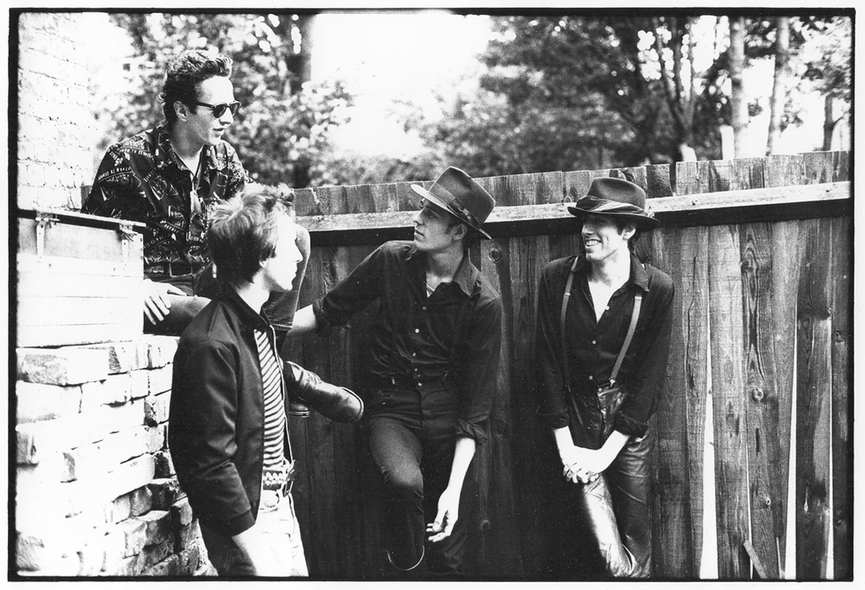 The Clash in a black and white photograph by Pennie Smith. They are in a back alley of a recording studio for the album 'London Calling' standing in front of a fence. They are casual & relaxed together, all smiling and enjoying being with one another.