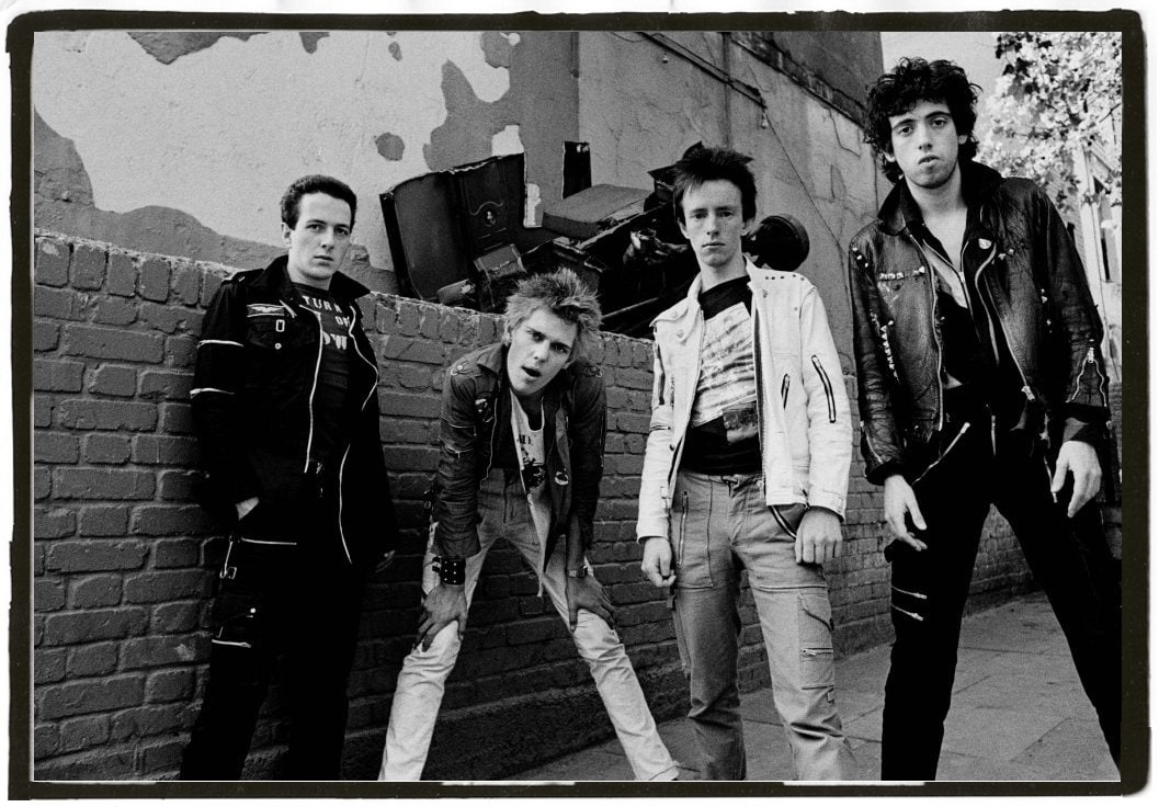 The Clash in a black and white image where they all are looking at the camera in a slight confrontational way. They are dressed in Punk attire of skinny pants & leather jackets.