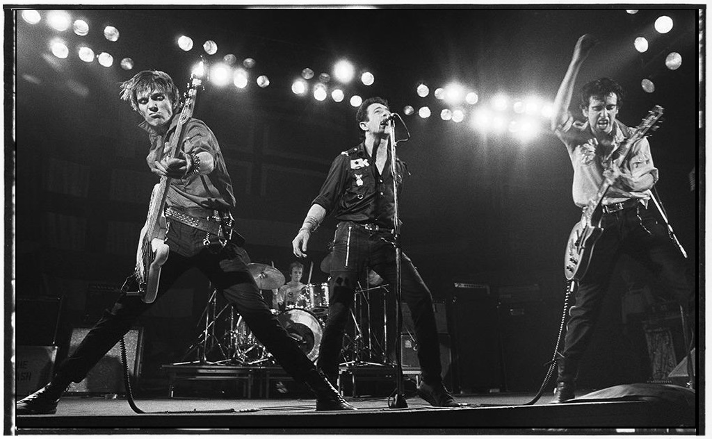 The Clash in a live photograph by Bob Gruen where their energy is on display. Guitarist & bassist Mick and Paul are captured in mid playing with fierce looks on their faces with Joe singing steady in the middle and drummer Topper behind.