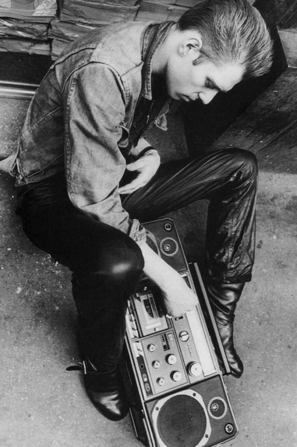 This black and white photograph shows Paul in an outfit of a jean jacket, leather pants, biker boots, pinkie ring, and a 1950s slicked back hairdo. He has a boombox to listen to music on the streets.