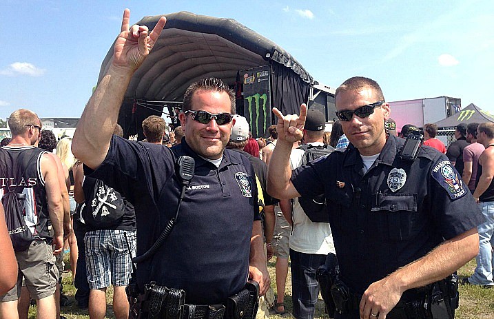 Two police men stand in front a warped tour stage while holding up the rock-on symbol with their hands