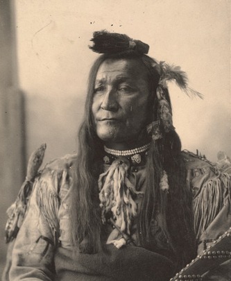 Chief Mountain, from the Blackfeet tribe in a sepia toned portrait from 1898. He is wearing traditional Native American clothing, which includes feathers in his hair.