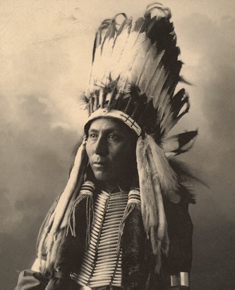 Native American Cheyenne Tribe Member Huggle Big Horse photographed in sepia tone in 1898 while wearing traditional clothing which includes a feather headdress.