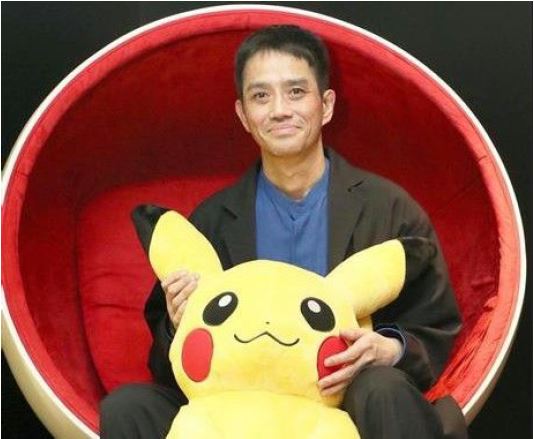 An image of Satoshi Tajiri, the creator of Pokemon. Tajiri is smiling at the camera, while he sits in a red circular chair and holds a large, yellow toy Pikachu.