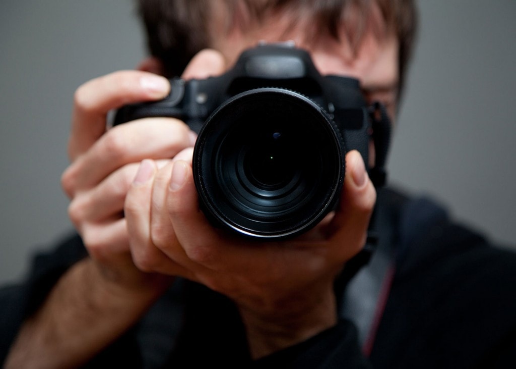 A photo of a black camera in the foreground, with the photographer, a man with his face hidden behind the camera, blurred in the background