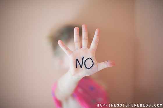 A nondestinct child in a pink shirt holding her hand up towards the camera to cover her face. The word NO is written across her palm.