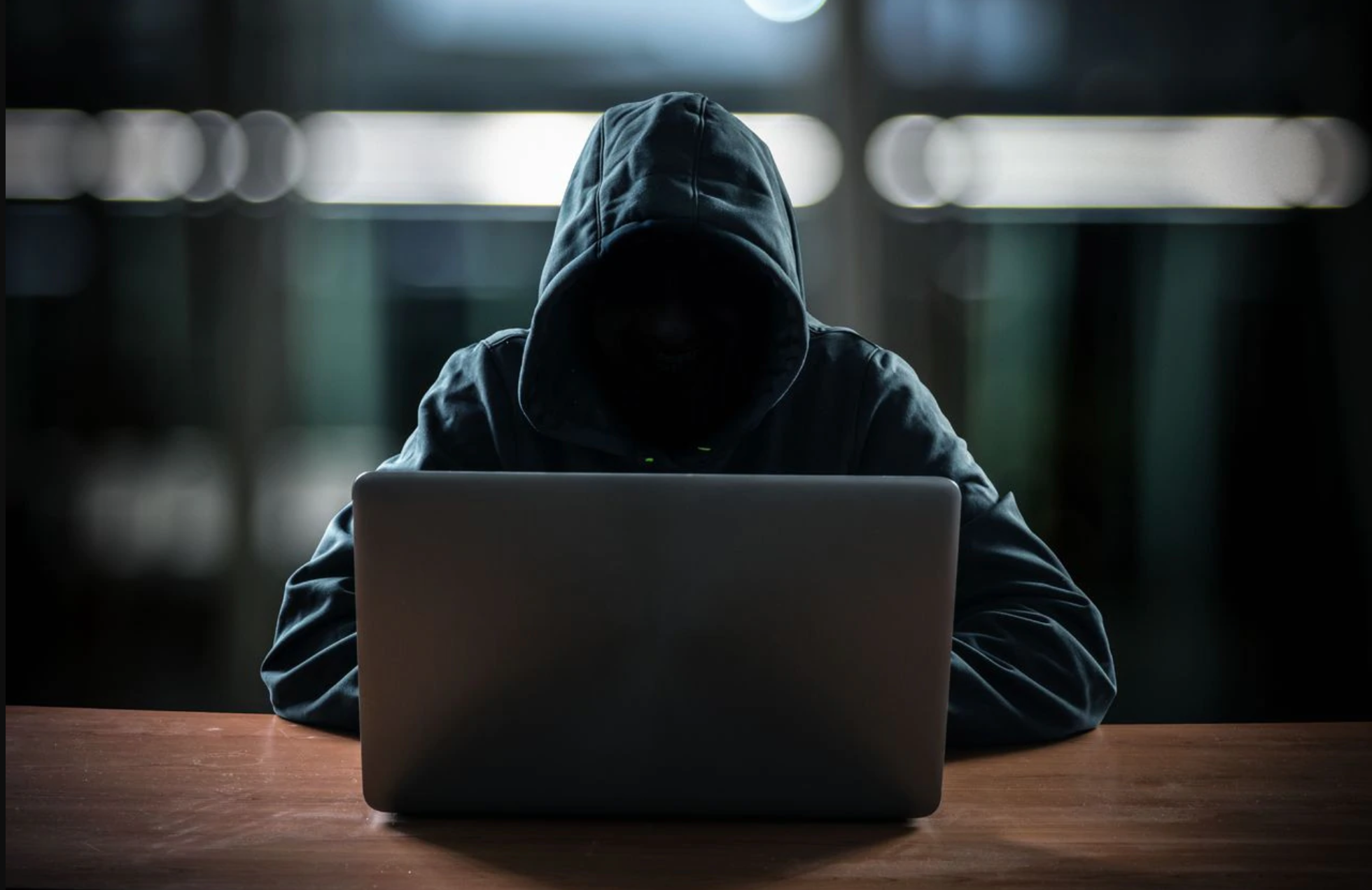 A man in a dark gray hoodie sits against a blurred background facing the camera. The hood of his hoodie is keeping his face dark and discreet. He is looking over a silver laptop.