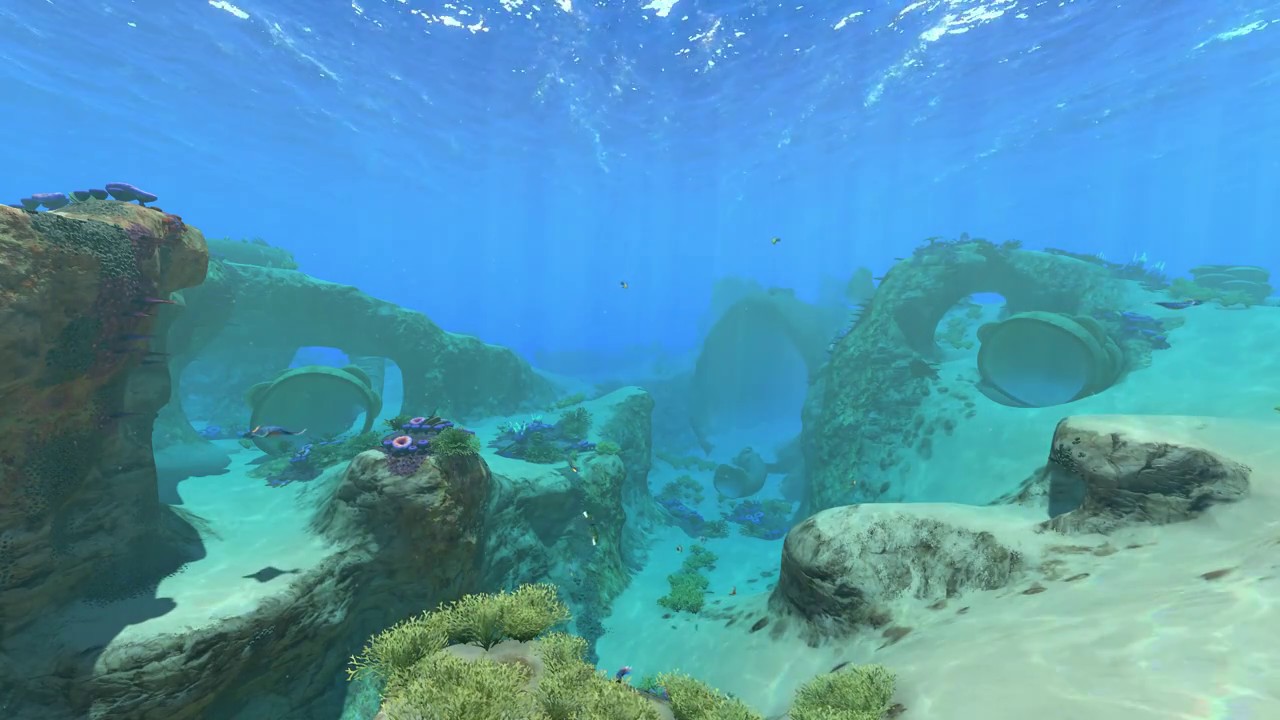 a screenshot of the safe shallows. The water is clear and blue, and the ground is soft and sandy. Jaggic rock makes up small cliffsides in the middle distance. A large tunnel can be seen in the far distance.