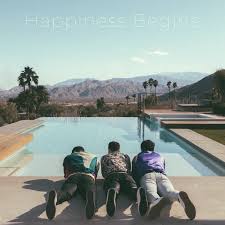 Album cover of three men lying on their stomachs at the edge of a modern pool, facing distant mountains and palm trees under a hazy sky, with Happiness Begins across the top.