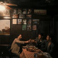 Dimly lit scene of three men seated around a checkered-tablecloth dinner table in a cozy room, raising glasses together beneath framed photos on the wall.