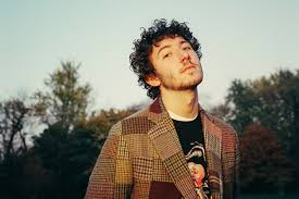 Outdoor portrait of a young man with curly hair wearing a patchwork-style blazer, standing in a field with trees behind him and tilting his head slightly.