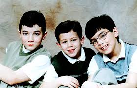 Childhood portrait of three young boys sitting close together and smiling at the camera against a soft studio backdrop.