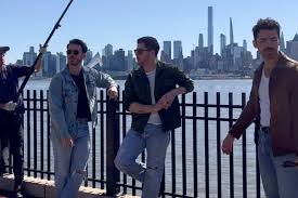 Behind-the-scenes style image of three men in casual jackets and jeans standing along a riverside railing, with a city skyline in the background and a crew member holding equipment to the side.
