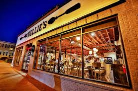 Exterior evening shot of a warmly lit restaurant with large front windows showing people dining inside and a sign across the top of the building that reads, Nellies.