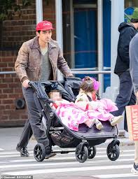 Street photo of a man in a red cap and brown jacket pushing a double stroller with two small children dressed in bright outfits while crossing an intersection.