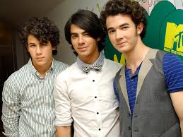 Three young men with curly hair stand close together at an event, two wearing collared shirts and one in a vest, looking toward the camera.