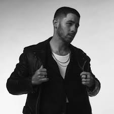 A man in a leather jacket and T‑shirt poses in black and white, holding the jacket lapels and gazing slightly to the side against a plain background.