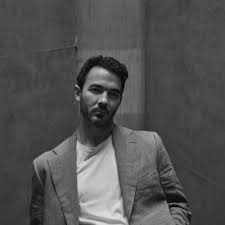 A man in a blazer and T‑shirt poses in black and white against a textured wall, looking toward the camera with a relaxed expression.