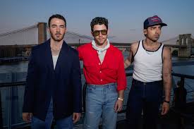 Three casually dressed men stand on a rooftop or balcony at dusk with a bridge in the background, one wearing sunglasses and another a baseball cap.