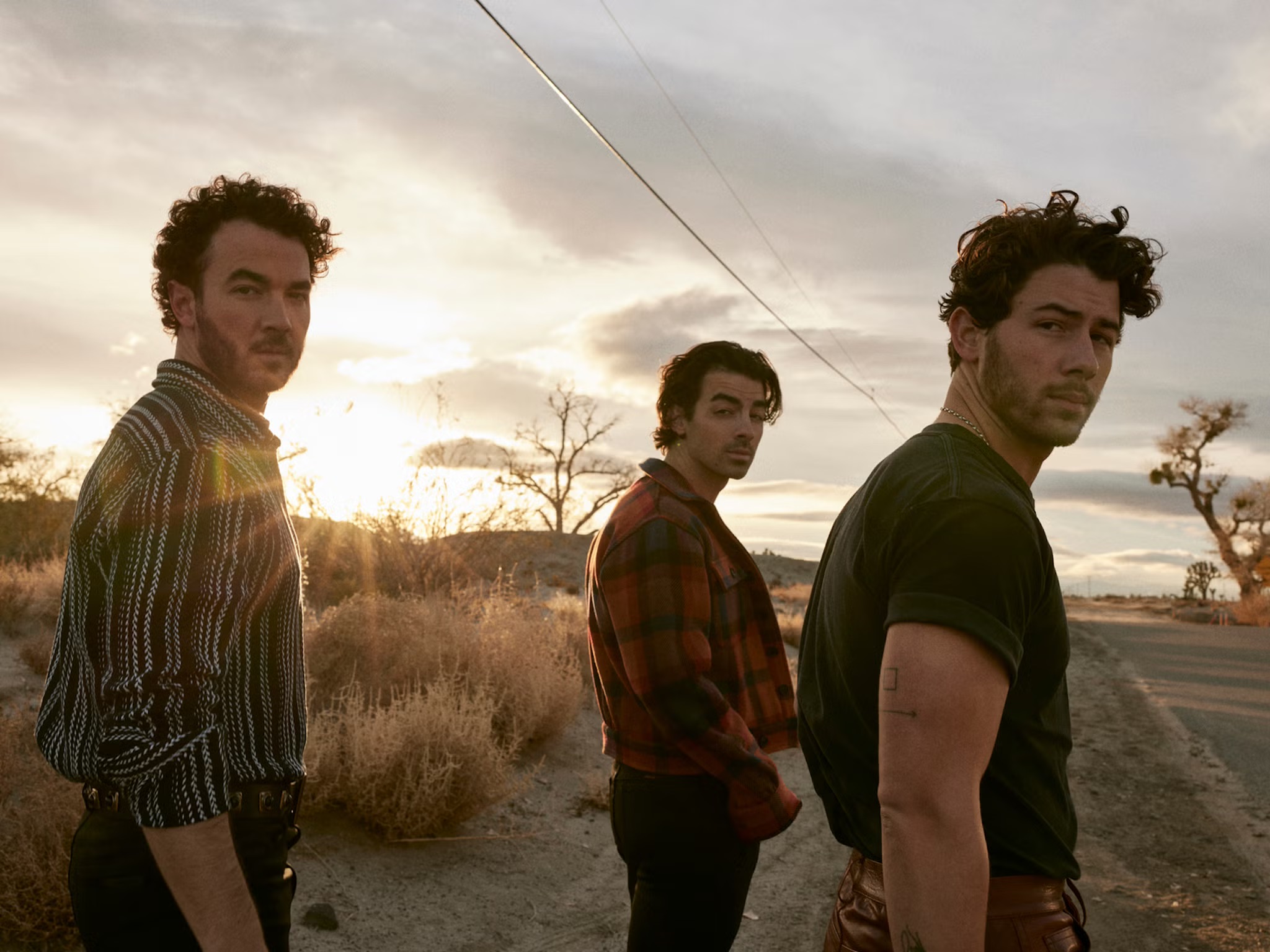 Three men walk along a dusty road at sunset, all turning their heads toward the camera with power lines and bushes stretching into the distance.