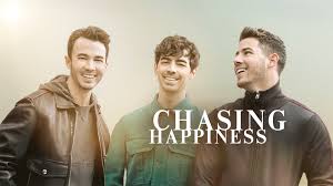 Promotional image with three smiling men standing side by side against a soft, hazy background, with the title “Chasing Happiness” in large white letters beside them.
