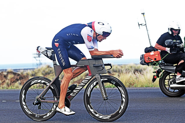 a man in a blue tri-suit biking on a road next to another person