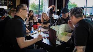 A group of six people sit around a black, rectangular table ready to play a game of Dungeons & Dragons. The camera points behind the Dungeon Master's screen. Behind the screen, we can see the player's dice, mats, and minis.