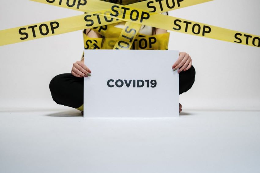 A person in black pants sitting on the floor of a white room holding a sign that says 'Covid19.' This person is wrapped up in black and yellow caution tape, but instead of 'caution,' the tape says 'stop.'