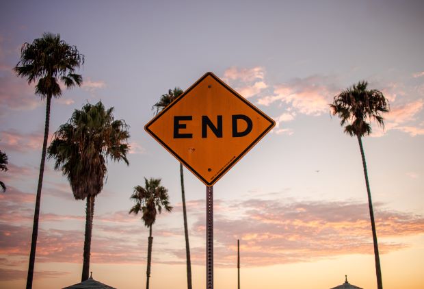 A yellow regulatory sign that says 'End' on it. Behind the sign, we see tall palm trees and a pink sunset sky with fluffy thin clouds.'