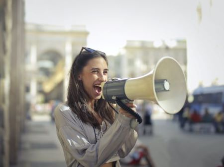 A brunette woman wearing white-toned clothes and with sunglasses on her head happily speaking into a white megaphone.