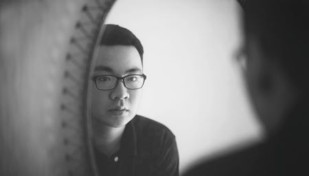 A man wearing a black shirt and glasses gazes at himself in a mirror with curiosity. The reflected face is in focus, but the rest of the image is blurred. The entire image is grayscaled.