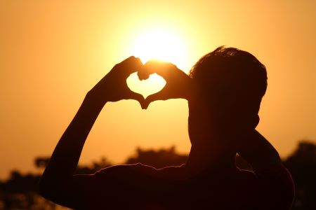 A man facing away from the camera towards a golden sunset. He holds his hands up in front of the sun and puts his hands in a heart shape. The sunlight pours through said heart.