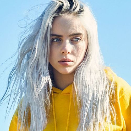 An image of Billie Eilish head-on, but she's not looking at the camera. She has blonde/grey hair and a yellow jacket on