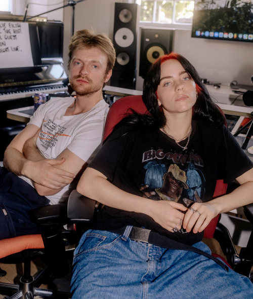 a picture of Billie Eilish with her brother Finneas sitting in their recording studio, which is Finneas's bedroom. They're both looking into the camera and not smiling. 