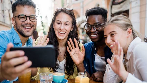 A group of four people gathered around a phone smiling and waving