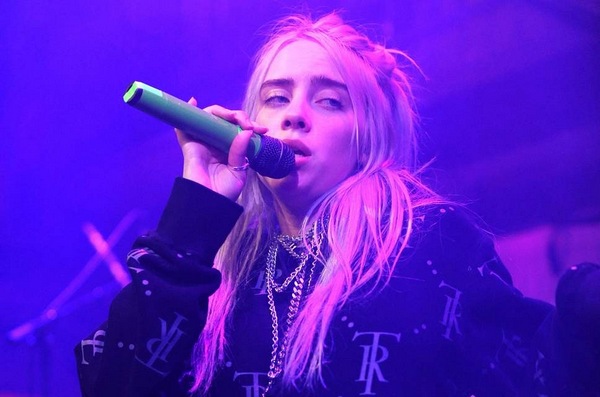 An image of Billie Eilish singing on stage. She's holding a microphone and looking out into the audience. Her mouth is slightly open, showing she's in the middle of singing