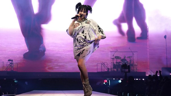 An image of Billie Eilish skipping on stage while she performs. She has dark hair with space buns. She has a white outfit on and in the background is the rest of the stage