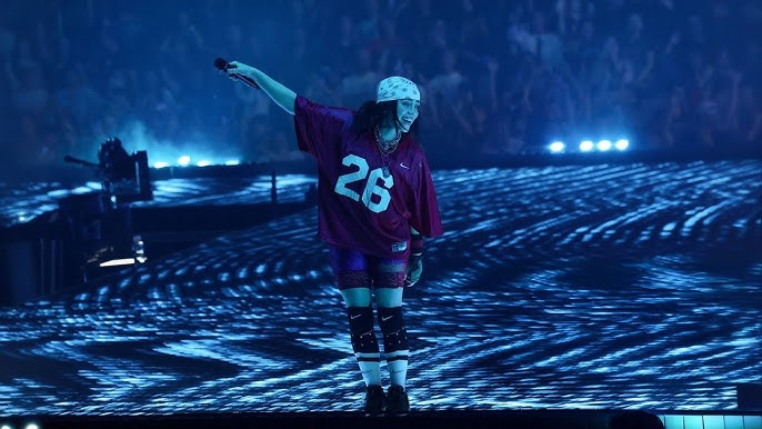 An image of Billie Eilish performing. She is holding her microphone out to the crowd and smiling. She has a white bandana on and a red outfit