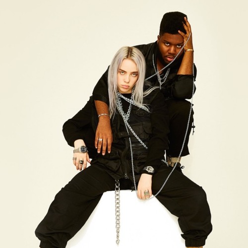 An image of Billie Eilish and Khalid. Billis is sitting in front of him. They're wearing all black and have chains wrapped around them