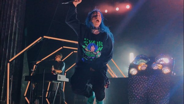 An image of Billie Eilish jumping up in the air on stage. She has blue hair and her bother Finneas is in the background on the keyboard