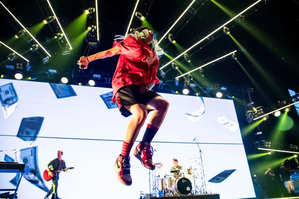 An image of Billie Eilish jumping on stage while performing. She's wearing red. Her brother Finneas is on the guitar in the background. The drummer is also in the background