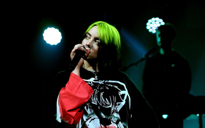 An image of Billie Eilish singing on stage. She's facing the camera and holding a microphone up to her face. She has green hair. Her brother Finneas can be seen in the background