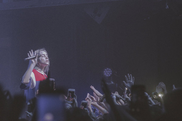 An image of Billie Eilish performing on stage. She is standing up, holding a microphone, and has her eyes closed. Hands of her fans are up in the air surrounding her