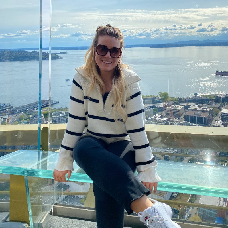 Carlee Linden wearing sunglasses and smiling while sitting on a glass bench with a oceanside city behind her and a beautiful view of an ocean inlet