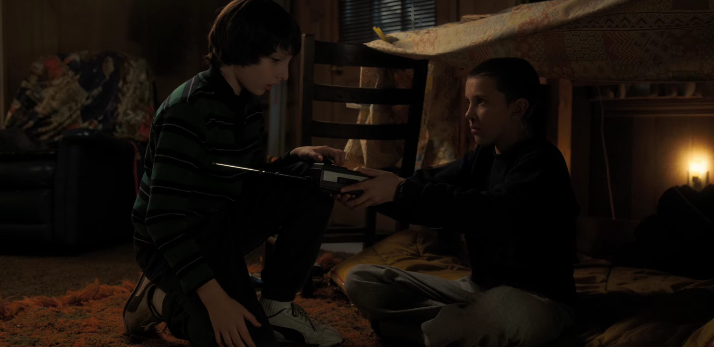 A young girl sits in a blanket fort. A young boy stands nearby talking to her and holding a hand-held radio.