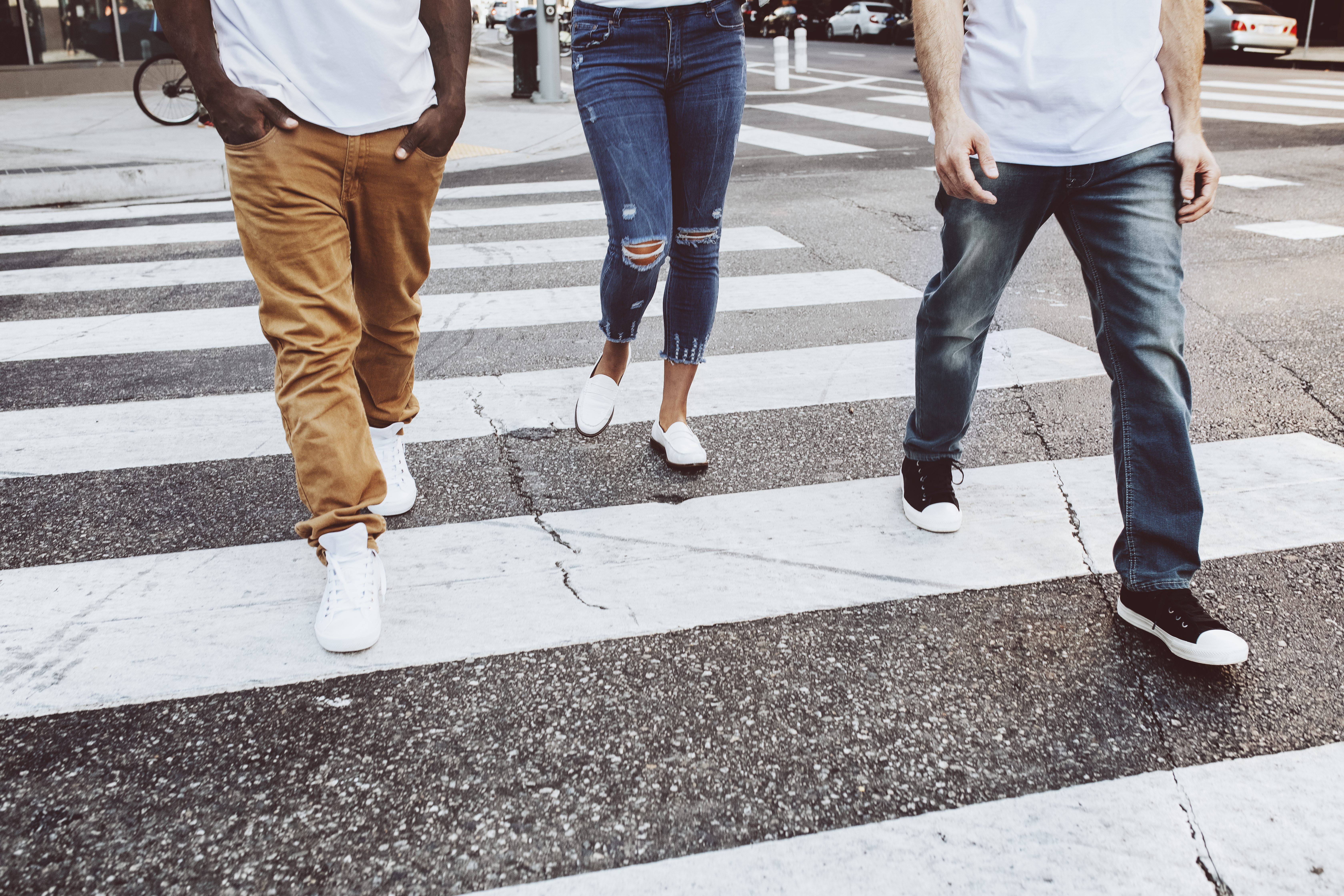 three people walking across a street on a white striped crosswalk, with only the bottom half of each person visible