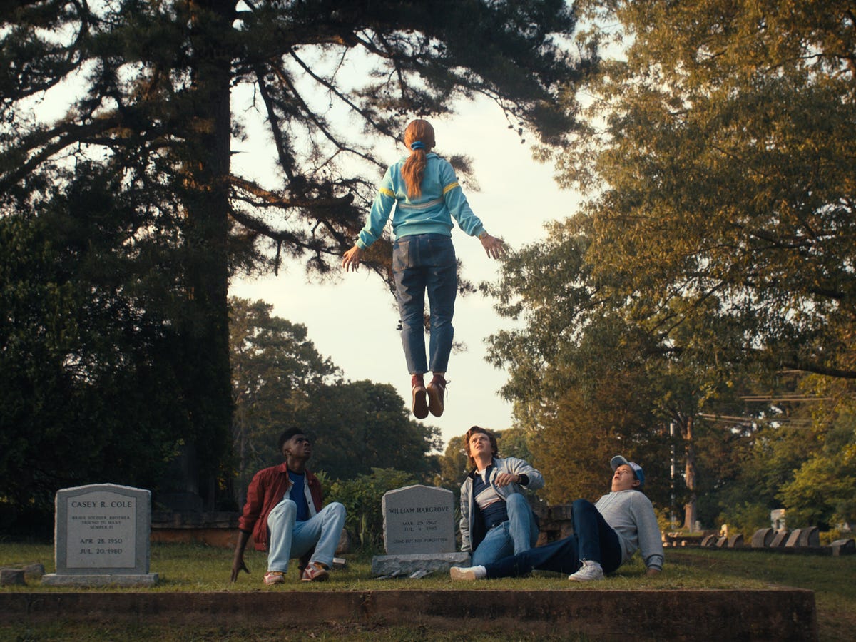 A girl rises up into the air in a cemetery with her arms outstretched while three boys look up at her in shock and worry.