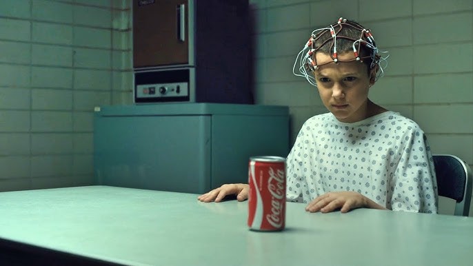 A young girl with a shaved head sits at a table focusing on a can of Coca-Cola in front of her. She is wearing a hospital gown and has a bunch of wires covering her head.