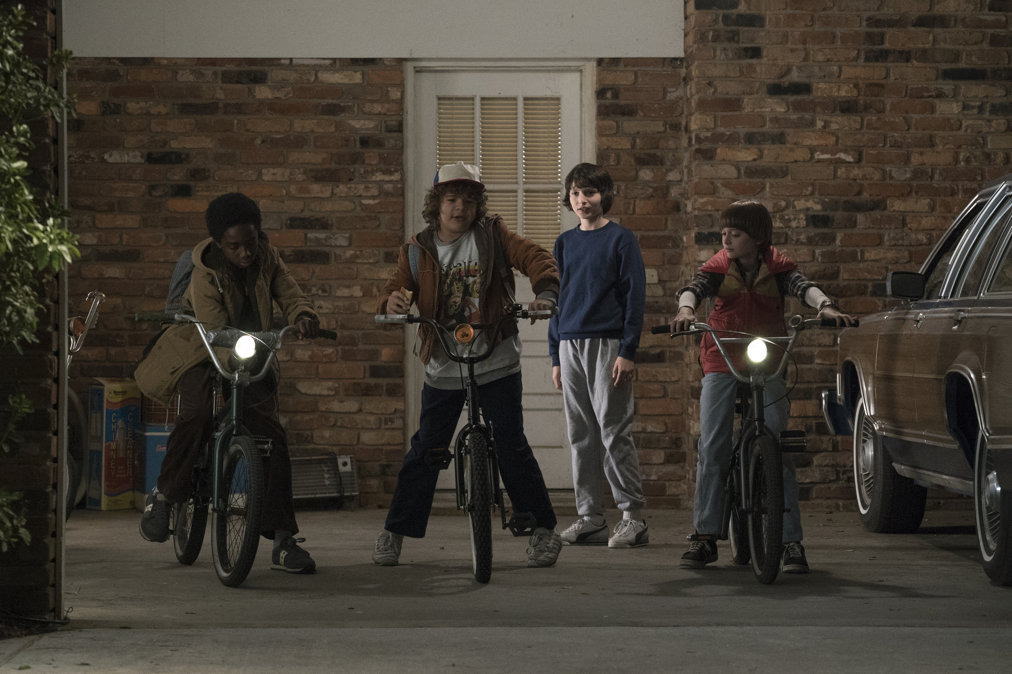 Three young boys climb onto their bikes while a fourth boy watches next to a house door.